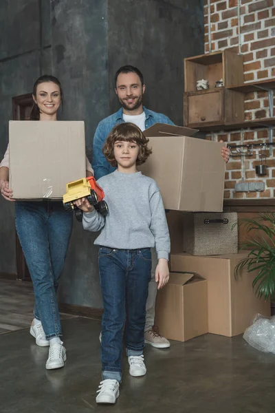 Familia feliz con cajas y juguete sonriendo a la cámara mientras se muda a casa - foto de stock