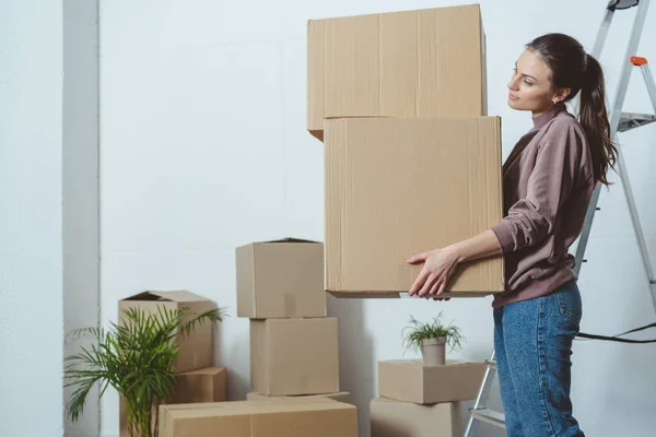 Vista lateral de la joven mujer sosteniendo pila de cajas mientras se muda a casa - foto de stock