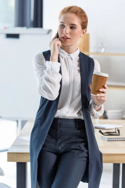 Woman with coffee cup in hands talks on smartphone in office — Stock Photo