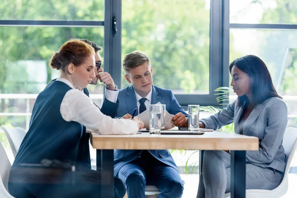 Detrás de la vista de cristal de hombres de negocios reflexivos y mujeres de negocios discutiendo proyecto en la oficina moderna - foto de stock