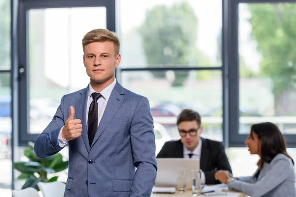 Jovem empresário mostrando polegar acima gesto na frente de seus colegas no escritório moderno — Fotografia de Stock