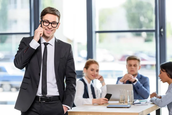 Homme d'affaires souriant parlant sur smartphone devant ses collègues dans un bureau moderne — Photo de stock