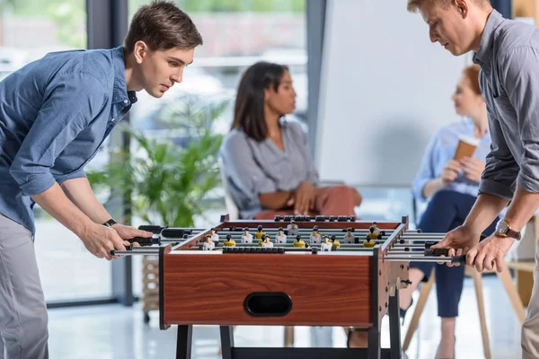 Dois homens de negócios jogando futebol de mesa, enquanto jovens empresárias conversando durante a pausa no escritório moderno — Fotografia de Stock
