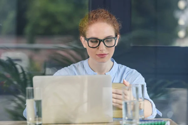 Behind the glass view of young businesswoman in glasses working by laptop — Stock Photo