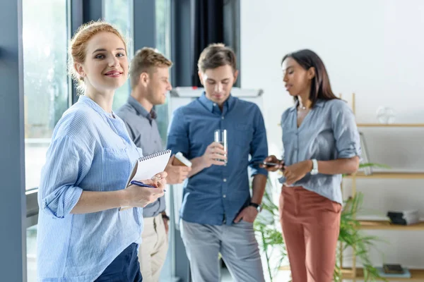 Jeune femme d'affaires avec bloc-notes devant ses collègues discutant du projet dans un bureau moderne — Photo de stock