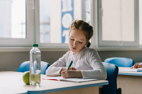 Focused little schoolgirl writing in classroom at school — Stock Photo