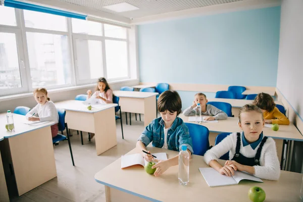 Groupe d'enfants avec des pommes et de l'eau assis en classe — Photo de stock