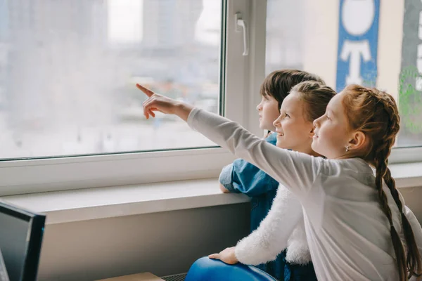 Colegiales adorables mirando a la ventana juntos en el aula y apuntando a algún lugar - foto de stock