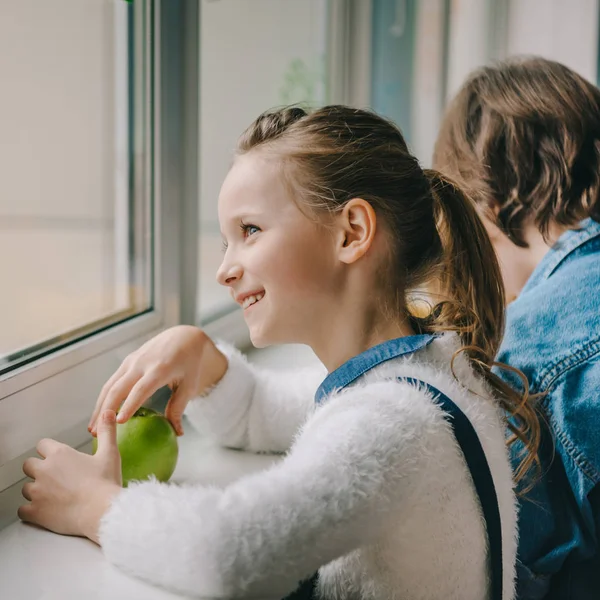 Hermosa colegiala con manzana fresca mirando a través de la ventana en el aula - foto de stock