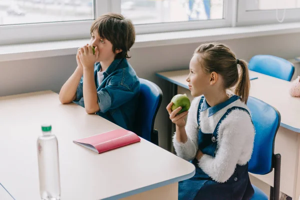 Compañeros de clase sentados en el escritorio y comiendo manzanas en el descanso - foto de stock