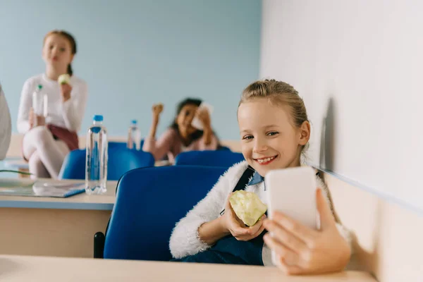 Feliz adolescente colegiala tomando selfie en aula mientras comer manzana - foto de stock