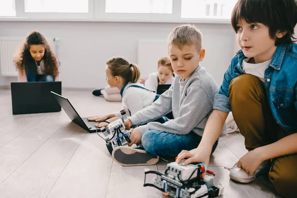 Fokussierte Kinder programmieren Roboter mit Laptops, während sie auf dem Boden sitzen, Stamm-Bildungskonzept — Stockfoto