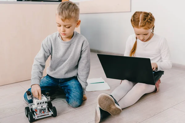 Konzentrierte Kinder sitzen mit Roboter und Laptop auf dem Boden — Stockfoto