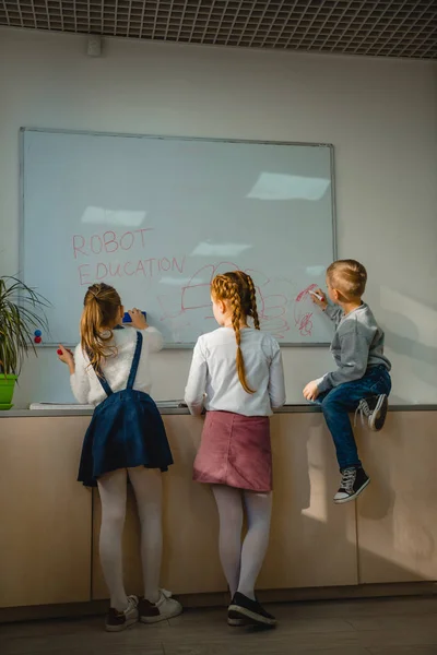 Vista posterior de los niños escribiendo y dibujando señales de educación robot en pizarra blanca - foto de stock