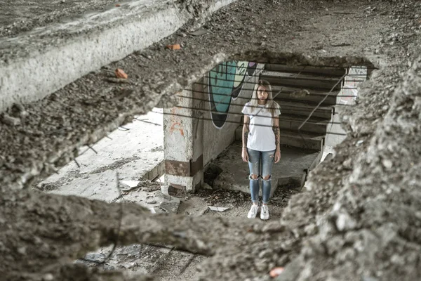 Nézd át a rostélyon. Egy magányos lány, egy fehér pólót és farmert áll egy romos ház romjai. Szomorú kifejezést, egy tragikus hangulatát. — Stock Fotó
