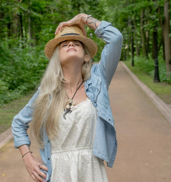 Leuke vrouw in lichte witte jurk en het dragen van een stro hoed poseren op camera in park buiten tegen groene achtergrond. Lange witte haren. — Stockfoto