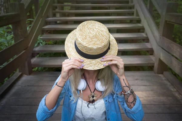 Linda jovem loira hipster mulher sentada nas escadas de madeira fecha o rosto chapéus de palha. A mulher está vestida com uma camisa jeans e vestidos de verão brancos . — Fotografia de Stock