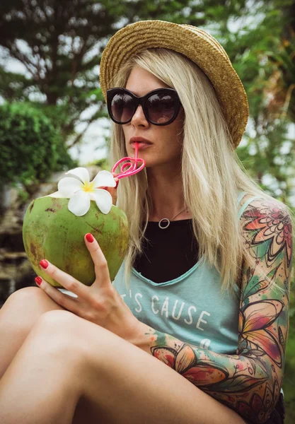 Vacker ung blond hipster i en stråhatt och solglasögon som sitter i en tropisk trädgård med kokos i hand. Regn helgen beach party — Stockfoto