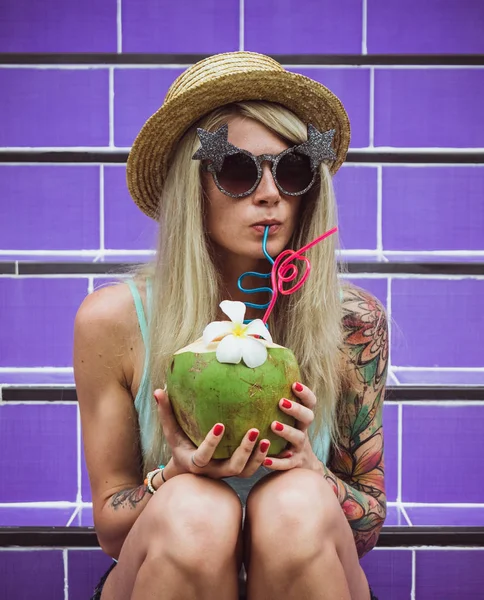 Belle jeune hipster blonde dans un chapeau de paille et des lunettes de soleil assis sur les escaliers violets avec de la noix de coco à la main. Week-end pluie plage fête — Photo