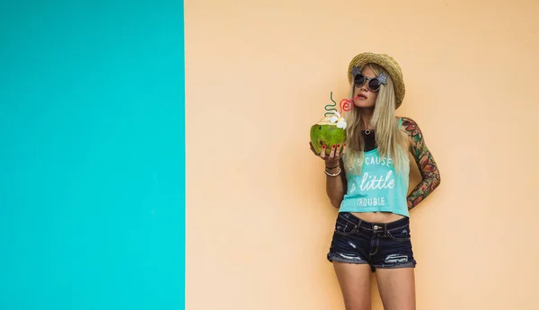Vacker ung blond hipster i denim shorts och en stråhatt, solglasögon står mitt i de färgglada väggarna med en kokosnöt i hand. Tropiska helg, beach party. — Stockfoto