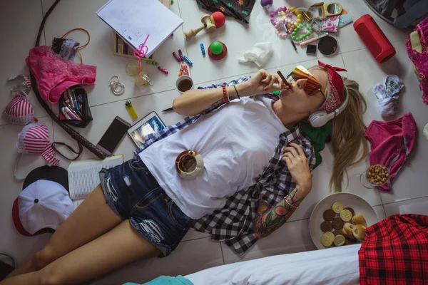 Na studeren blonde student, liggend op de vloer in de hoofdtelefoons en zonnebril, luisteren naar muziek en het roken van een sigaret. Het beeld van een moderne student, onderwijs, accessoires van een moderne jonge meisje — Stockfoto