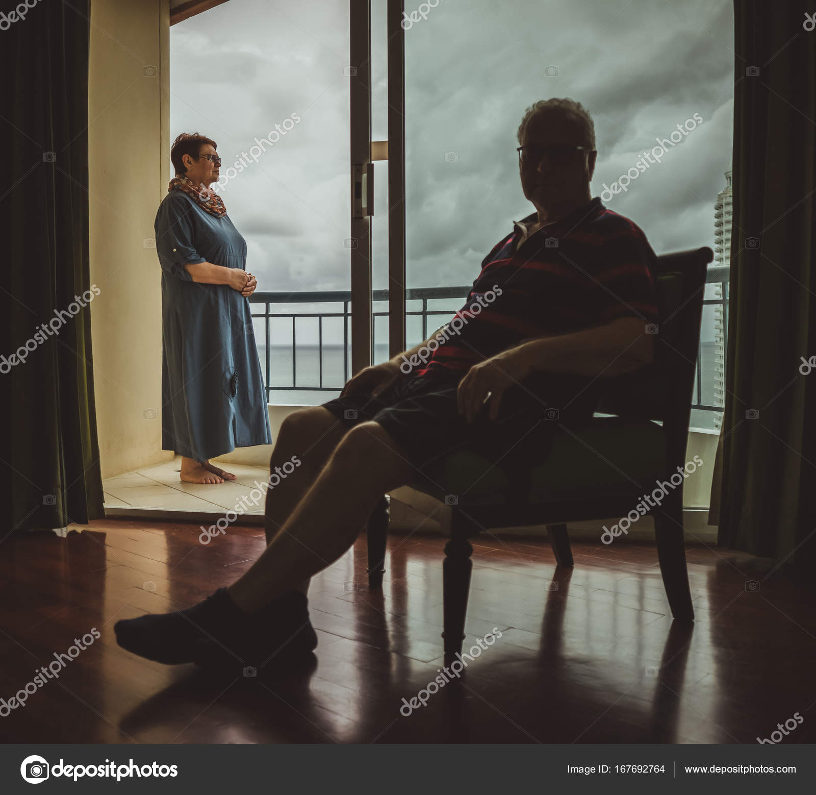 An Elderly Married Couple At Home The Husband Sits Indoors