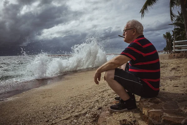 Пожилой человек в очках и в полосатой футболке, сидящий на пляже и смотрящий на шторм курит сигару. Мудрый человек в возрасте, тема одиночества . — стоковое фото