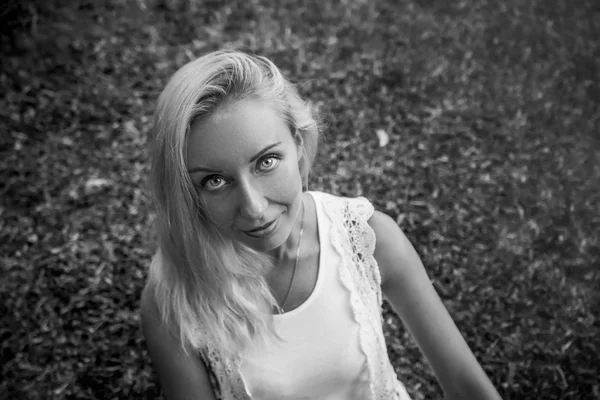 Güzel genç sarışın kadın yeşil çimenlerin üzerinde oturur ve kameraya güzel gri gözleri ile görünüyor. Modern yaşam tarzı ve gevşeme kavramları. Siyah ve beyaz.