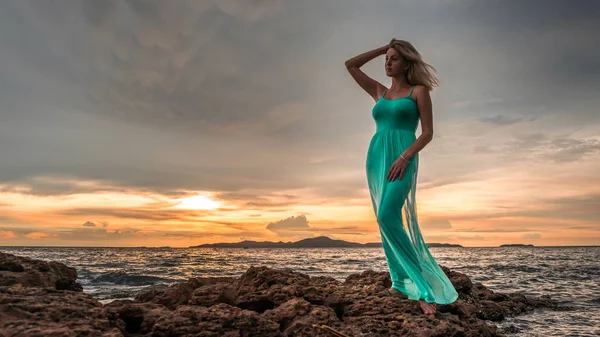 En vacker sexig blondin i en lång turkos klänning står på havet mot en bakgrund av en ljusa solnedgång — Stockfoto