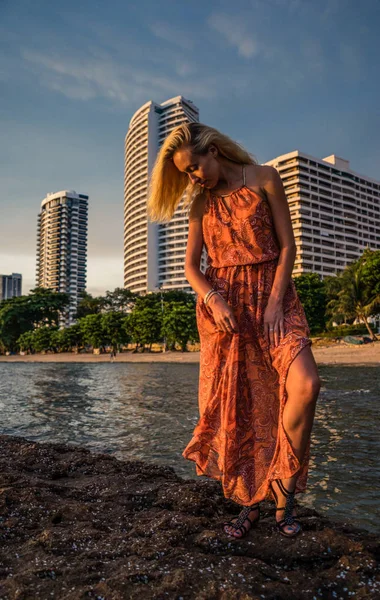 Блондинка в светло-красном платье стоит у моря на заднем плане курорта поздним полуденным солнечным светом. Поза для камеры — стоковое фото