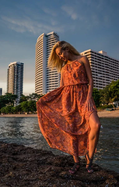 Beautiful blonde in a light red dress stands by the sea on the backdrop luxurious resort in the late afternoon sunlight. Posing for the camera — Stock Photo, Image