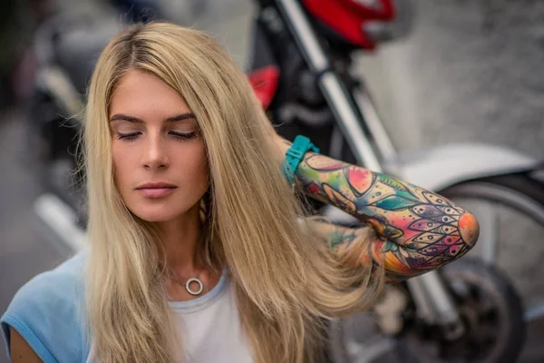 Portrait of a young blonde girl sitting on the background of the motorcycle. Womens hands with a tattoo. Urban fashion. — Stock Photo, Image