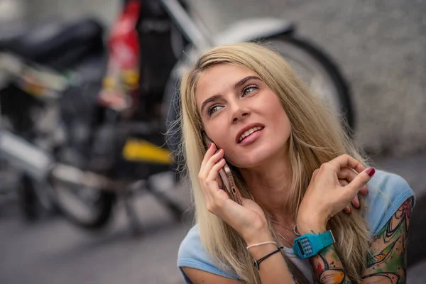 Portrait of young blonde sitting on the background of the motorcycle. Talking on the phone. Female hands with a tattoo. Urban fashion. — Stock Photo, Image