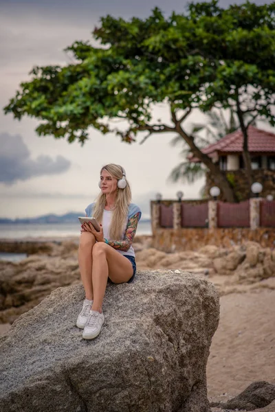 Gyönyörű szőke nő ült a szikla a tengerparton, és hallgató-hoz zene, vagy a tabletta. kezét a tetoválás. Stílus nyaralás. Modern lány — Stock Fotó