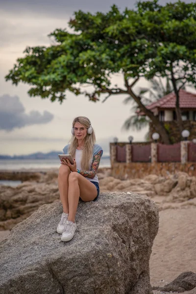 Красивая блондинка сидит на скале у моря и слушает музыку с планшета. руки с татуировками. Стильный летний отпуск. Современная девушка — стоковое фото