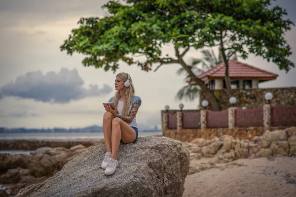 Mulher loira bonita sentada em um rock junto ao mar e ouvindo música com seu tablet. mãos com tatuagens. Estilo férias de verão. Menina moderna — Fotografia de Stock