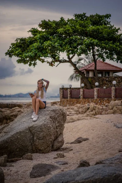 Beautiful blonde woman sitting on a rock by the sea and listening to music with your tablet. hands with tattoos. Style summer vacation. Modern girl — Stock Photo, Image