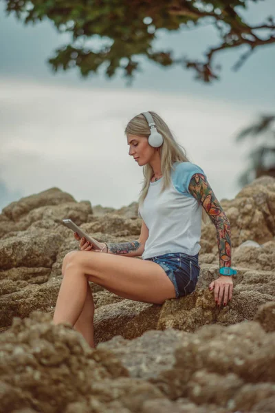 Красивая блондинка сидит на скале у моря и слушает музыку с планшета. руки с татуировками. Стильный летний отпуск. Современная девушка — стоковое фото