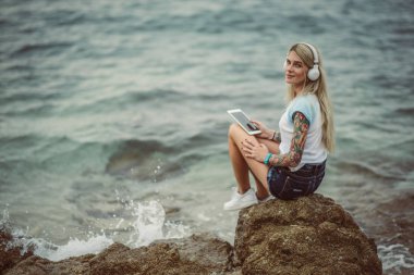 Bir kaya denizin kenarında oturan ve tablette şık kulaklıklar müzik dinlemek güzel sarışın bir kadın. elle bir dövme. Yaz tatili stili. Modern kız