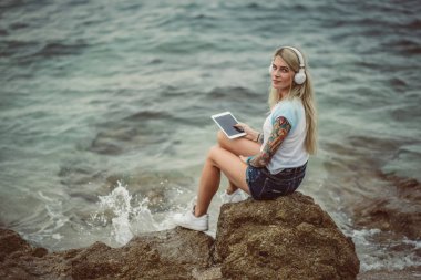 Bir kaya denizin kenarında oturan ve tablette şık kulaklıklar müzik dinlemek güzel sarışın bir kadın. elle bir dövme. Yaz tatili stili. Modern kız
