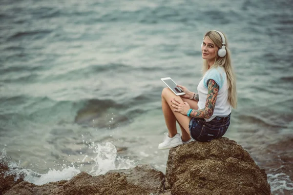 Mulher loira bonita sentada em uma rocha junto ao mar e ouvindo música em fones de ouvido elegantes no tablet. mãos com uma tatuagem. O estilo das férias de verão. Menina moderna — Fotografia de Stock