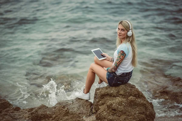 Bir kaya denizin kenarında oturan ve tablette şık kulaklıklar müzik dinlemek güzel sarışın bir kadın. elle bir dövme. Yaz tatili stili. Modern kız