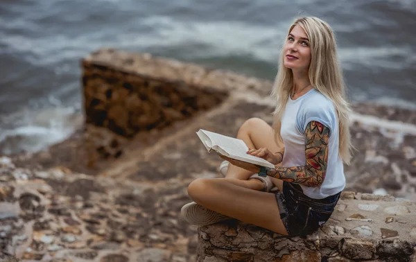 Mulher loira bonita sentada em um dique de pedra e lê livro. Mãos tatuadas. moderno, moderno, estudante — Fotografia de Stock