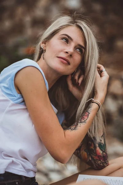 Красивая блондинка сидит на каменной набережной и читает книгу. Татуировка на руках. современный, модный, студент. Портрет крупного плана — стоковое фото
