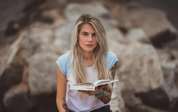 Красивая блондинка сидит на каменной набережной и читает книгу. Татуировка на руках. современный, трепетный, студенческий — стоковое фото