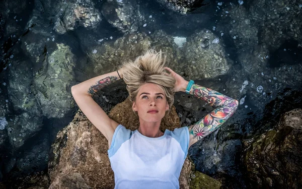 Portret van een mooie blonde die op de steen in de buurt van de zee ligt. Womens handen met een tatoeage. Moderne mode meisje — Stockfoto