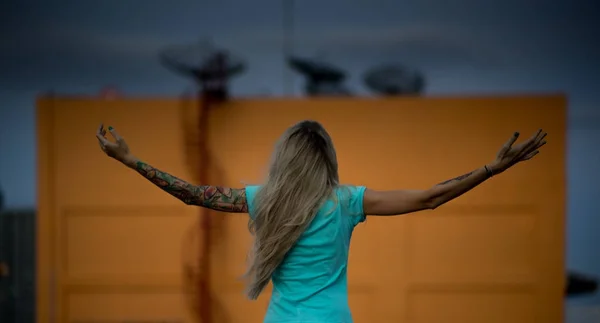 A hosszú, szőke haja áll háttal a kamera emelt karokkal a háttér narancssárga falak, nagy antennákkal. Türkiz pólót visel. Tetovált fegyverek. — Stock Fotó