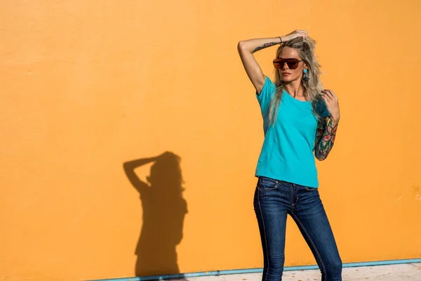 Mooie blonde in zonnebril corrigeert luxe haren. Portret op de achtergrond van de heldere oranje muur. Moderne hipster meisje met tatoeages. — Stockfoto