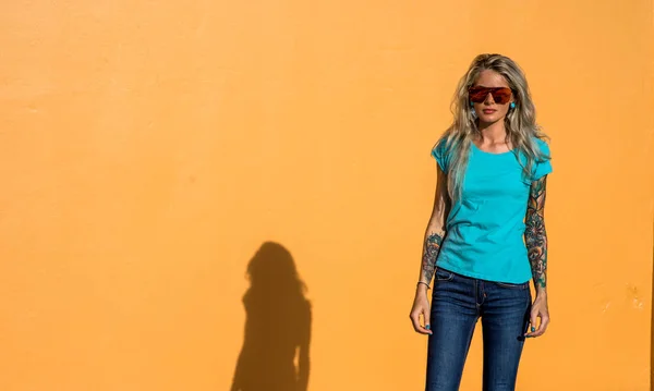 Mooie blonde in zonnebril kijken naar de camera. Portret op de achtergrond van de heldere oranje muur. Moderne hipster meisje met tatoeages. — Stockfoto