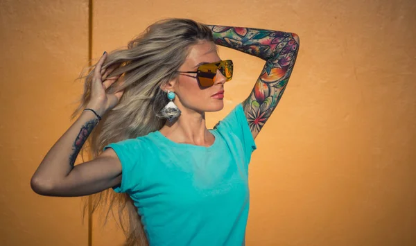 Mooie blonde in zonnebril poseren op de camera. Portret op de achtergrond van de heldere oranje muur. Moderne hipster meisje met tatoeages. Closeup portret. — Stockfoto